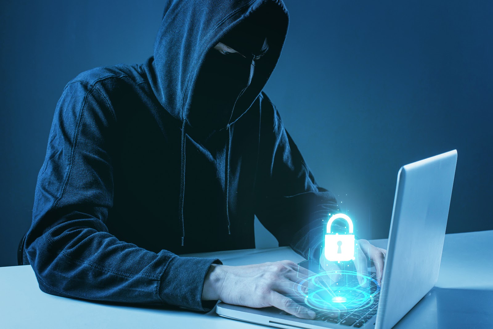 Persona encapuchada frente a un computador portátil con un candado digital sobre el teclado, representando un ciberataque o intento de vulnerar la seguridad informática.